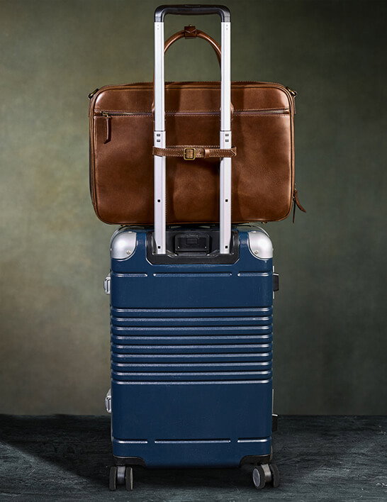 A tan leather briefcase with a buckle luggage strap sits on top of a navy blue hardshell spinner carry-on suitcase with a silver aluminum frame and extended telescoping handle, photographed against a dark olive green background.