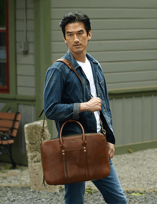 A man walks outdoors near a green-sided building, carrying a structured tan leather briefcase by its top handles with a matching shoulder strap over one hand. He wears a denim jacket over a white t-shirt and light wash jeans.
