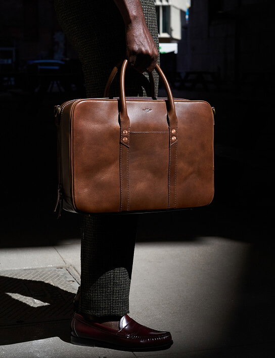 A man holds a structured tan leather briefcase by its dual top handles at his side, dramatically lit against a dark urban background. He wears dark green tweed trousers and burgundy penny loafers. The bag features a small logo emblem, copper rivets, and contrast stitching.