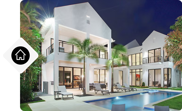 Two-story home with lit patio, lounge chairs, and a rectangular pool at night.