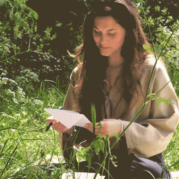 A woman sits among leafy greenery, opening and reading a greeting card outdoors.
