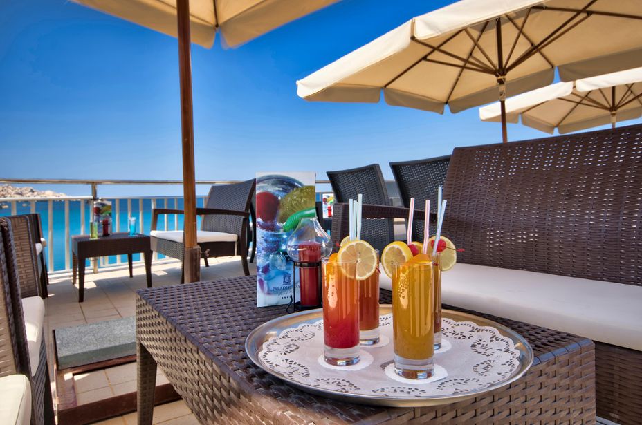 Three colourful cocktails with lemon slices on a silver tray atop a wicker table at a sunny seaside terrace with parasols and blue sea view