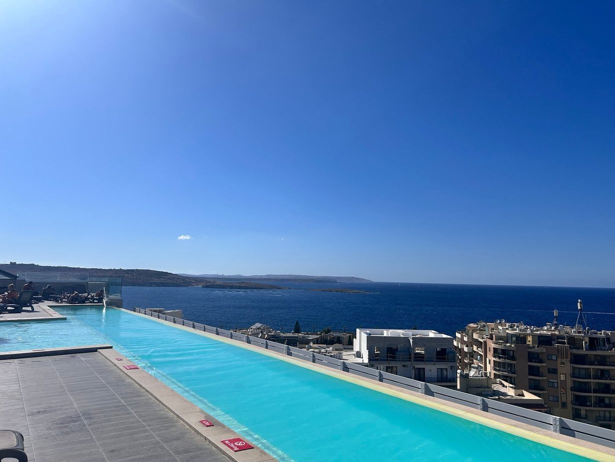 Rooftop infinity pool with turquoise water overlooking the Mediterranean Sea and coastal apartment buildings under a clear blue sky