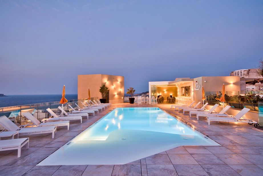 Rooftop hotel pool at dusk with illuminated infinity-style pool, rows of white sun loungers and sea view