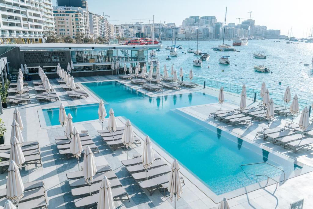 Rooftop hotel infinity pool with rows of sun loungers and closed umbrellas, overlooking a sunlit marina with boats and modern coastal buildings