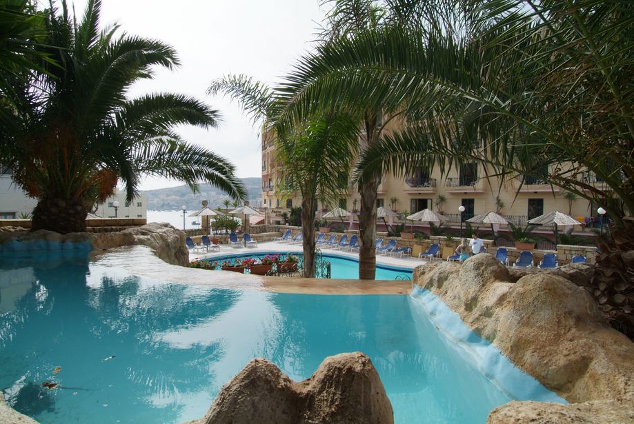 Seaside hotel resort pool with palm trees, rock features, sun loungers and parasols beside hotel and distant sea view