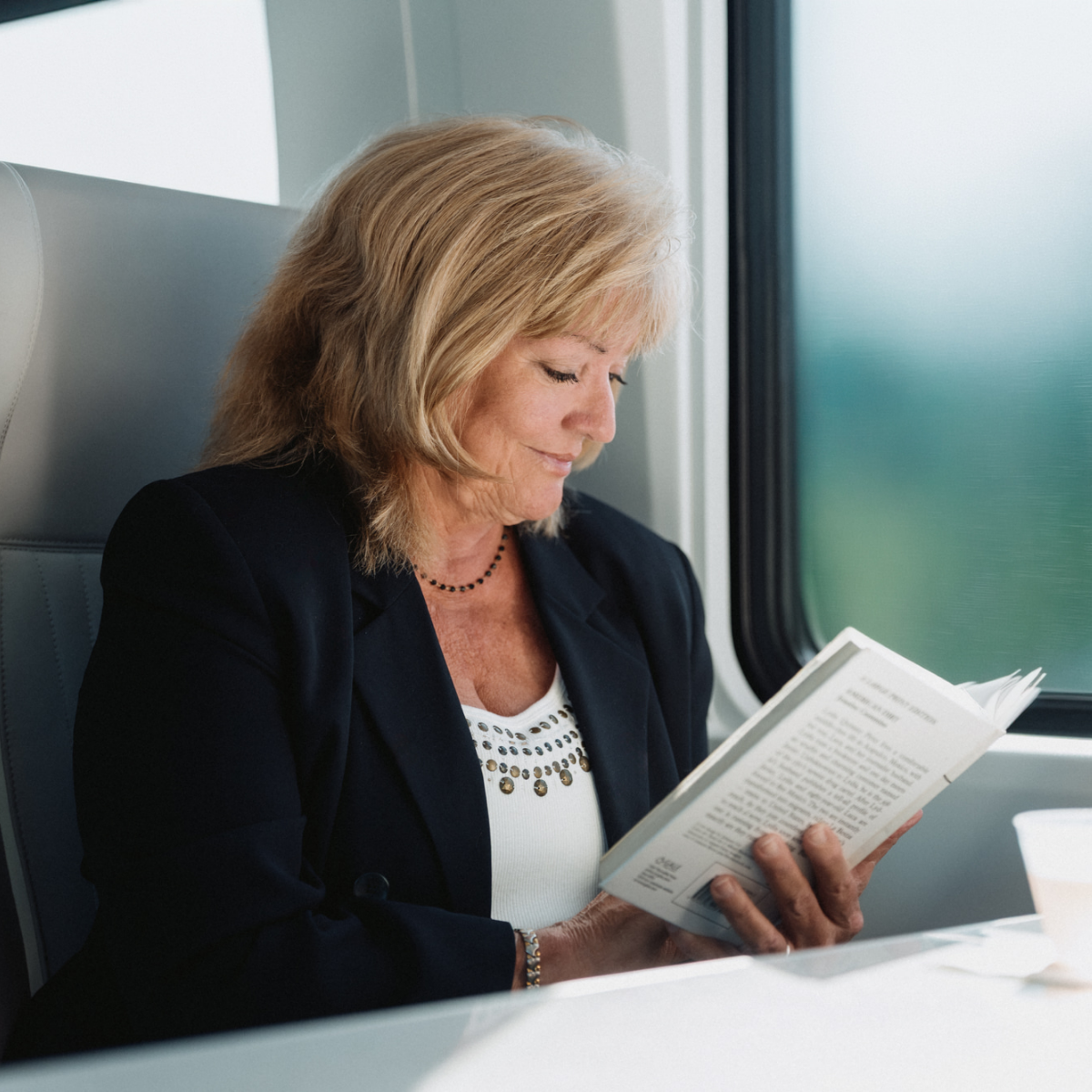 A mature woman sits by a window reading a book while traveling on a train. She wears a dark blazer over a white top and appears focused on the text. Soft daylight comes through the large window, illuminating her face and the pages. A cup is visible on the table in front of her, adding to the relaxed travel atmosphere.