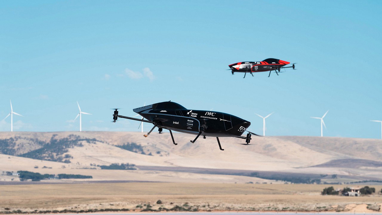 Two flying drones above a landscape with wind turbines in the background.
