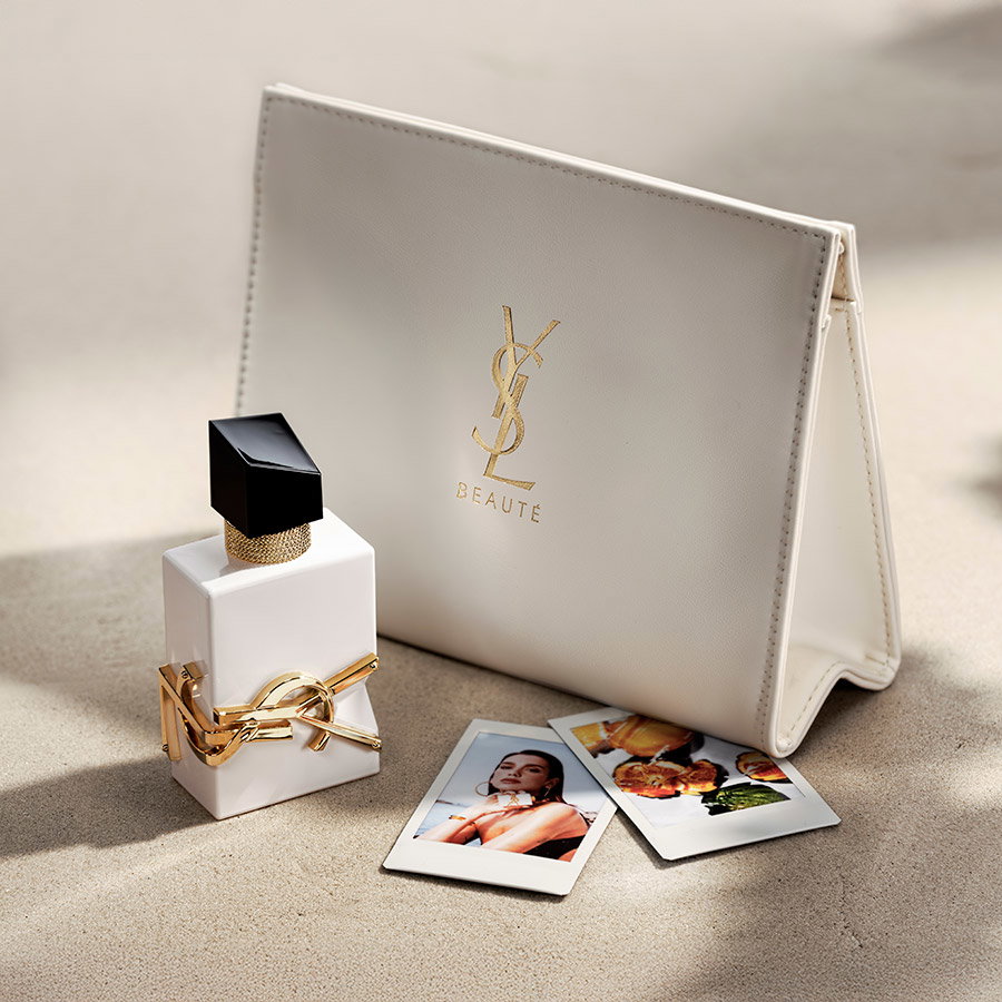 A white YSL Libre fragrance bottle displayed next to a matching white leather clutch and polaroid photos on sand.