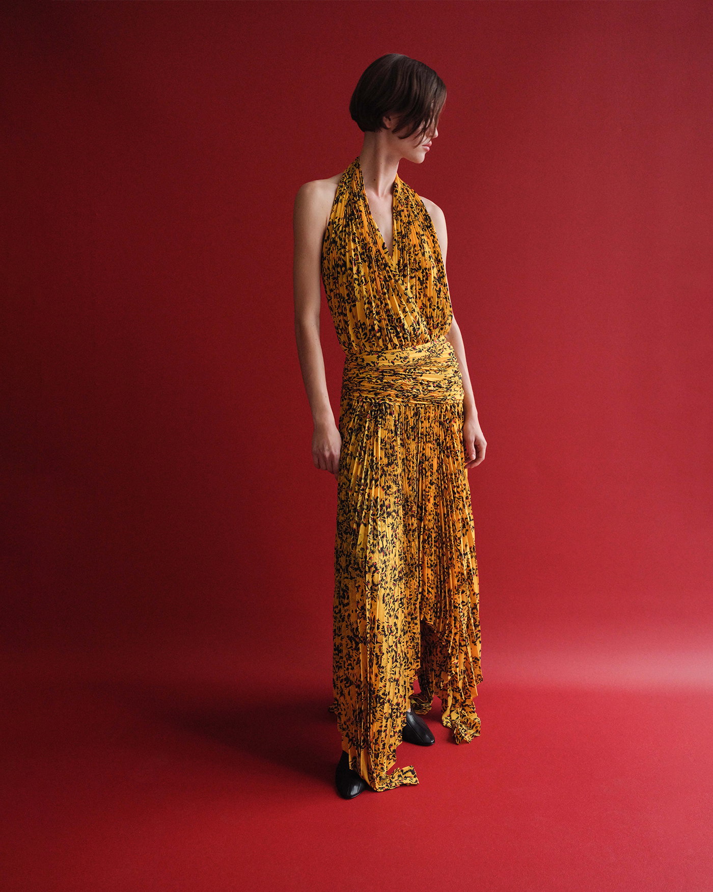 Image of model standing against a red seamless, wearing the Paulina Dress in Pleated Georgette in Gold Multi with the Tee 85 Pieced Pumps in Black