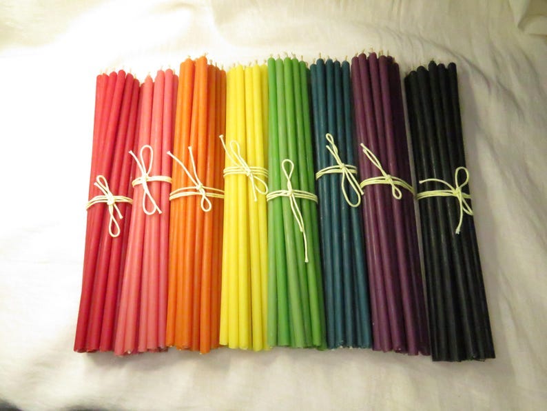 May include: Bundles of colorful taper candles in shades of red, pink, orange, yellow, green, blue, purple, and black. Each bundle is tied with a white string. The candles are arranged in a row, creating a vibrant display.