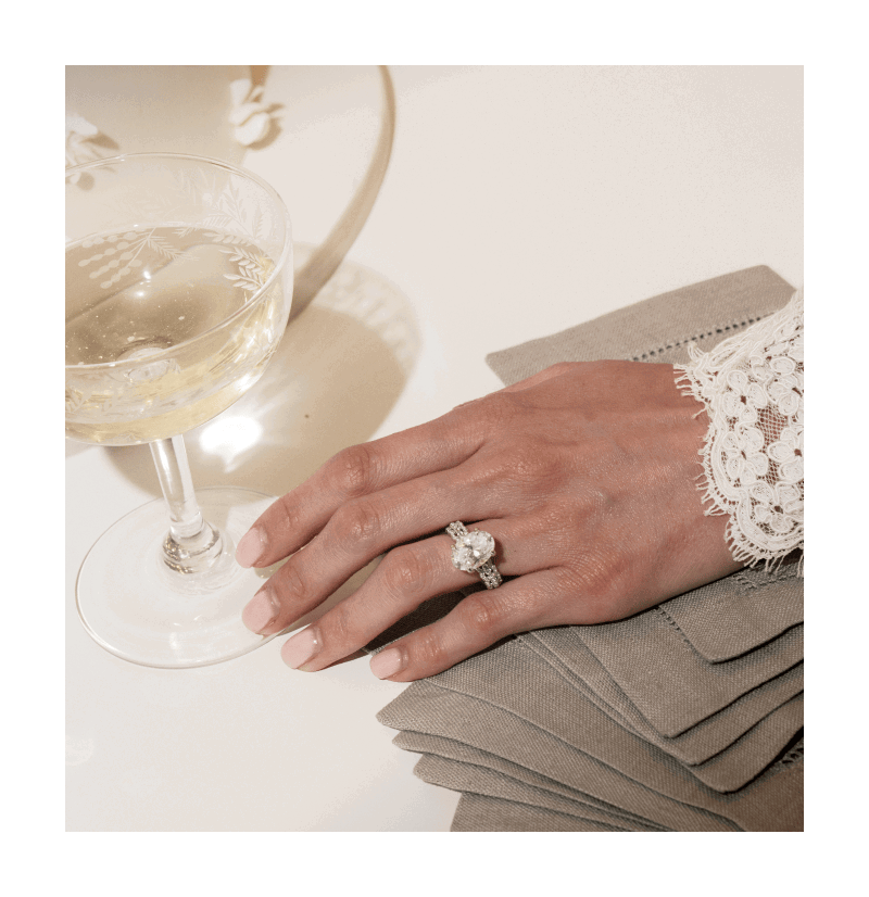 A hand wearing a ring with an oval cut diamond next to a glass of champagne. 