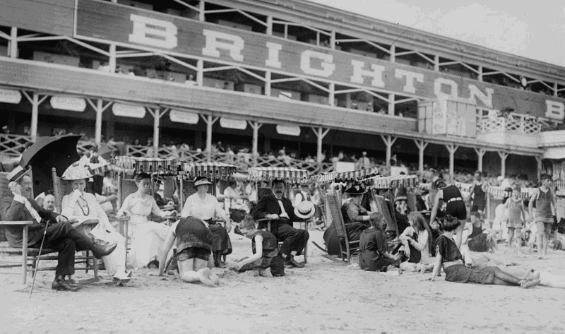 Black and white photo of people sitting and lounging on the sand in front of a large building labeled "Brighton Beach.