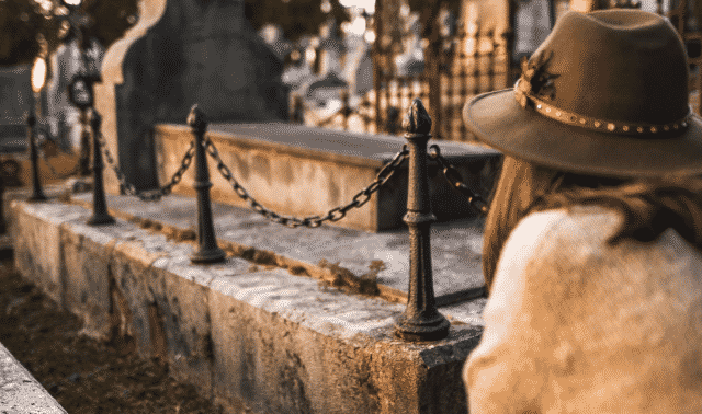 A person wearing a hat and a light-colored sweater is kneeling by a tombstone in a cemetery. The chain fence around the tombstone is visible.