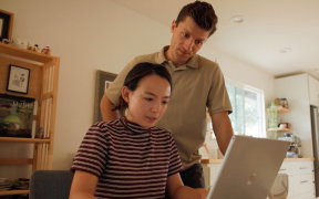 a woman looking at a laptop screen with a man standing behind her and looking over her shoulder