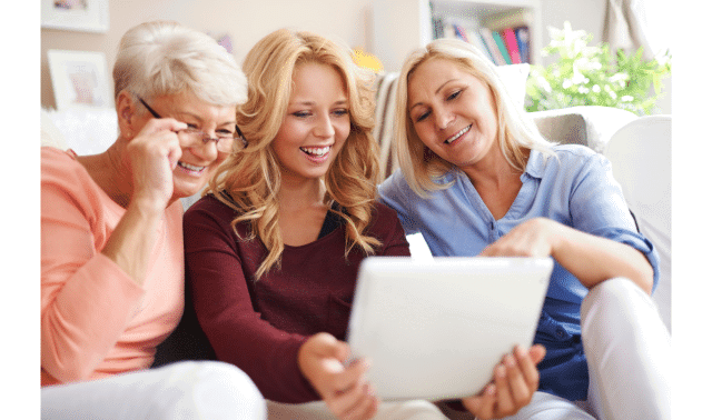Three women smiling and looking at a tablet while sitting on a sofa in a bright room.