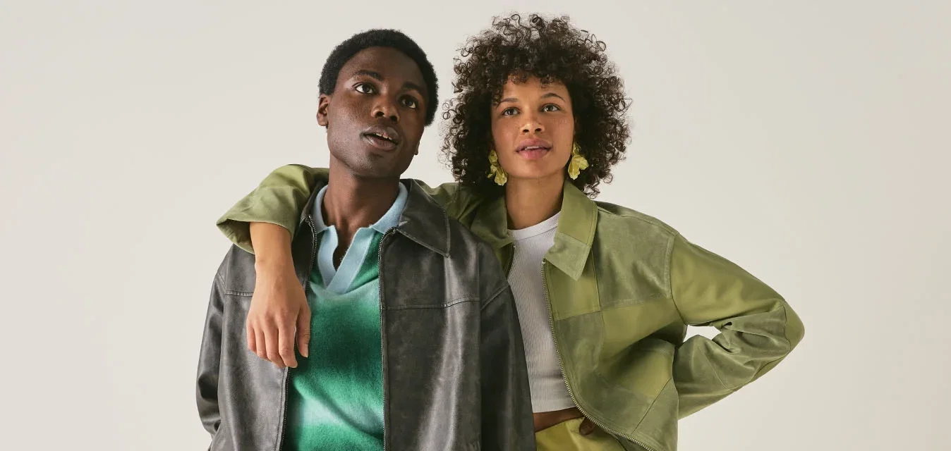 Due giovani adulti stanno vicini; uno indossa una giacca grigia e un maglione verde, l'altro una giacca verde chiaro e una gonna, posando davanti a uno sfondo semplice.
