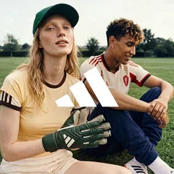 Due giovani in abbigliamento sportivo seduti sull’erba, uno indossa guanti da portiere e un cappellino, l’altro una maglia da calcio, all’aperto.