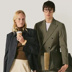 Uomo e donna in piedi, entrambi vestiti in abbigliamento smart casual; la donna tiene in mano una tazza di caffè e una borsa, l'uomo tiene una grande borsa di pelle.