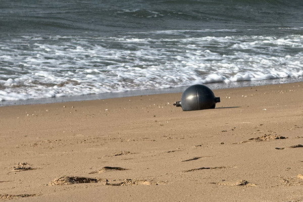 A grey sphere shines in the sun on the sand of a beach, with the sea in the background.