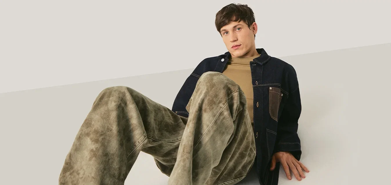 Jonge man zittend op de grond met gebogen benen, gekleed in losse groene broek, beige shirt en donker jasje tegen een neutrale achtergrond.