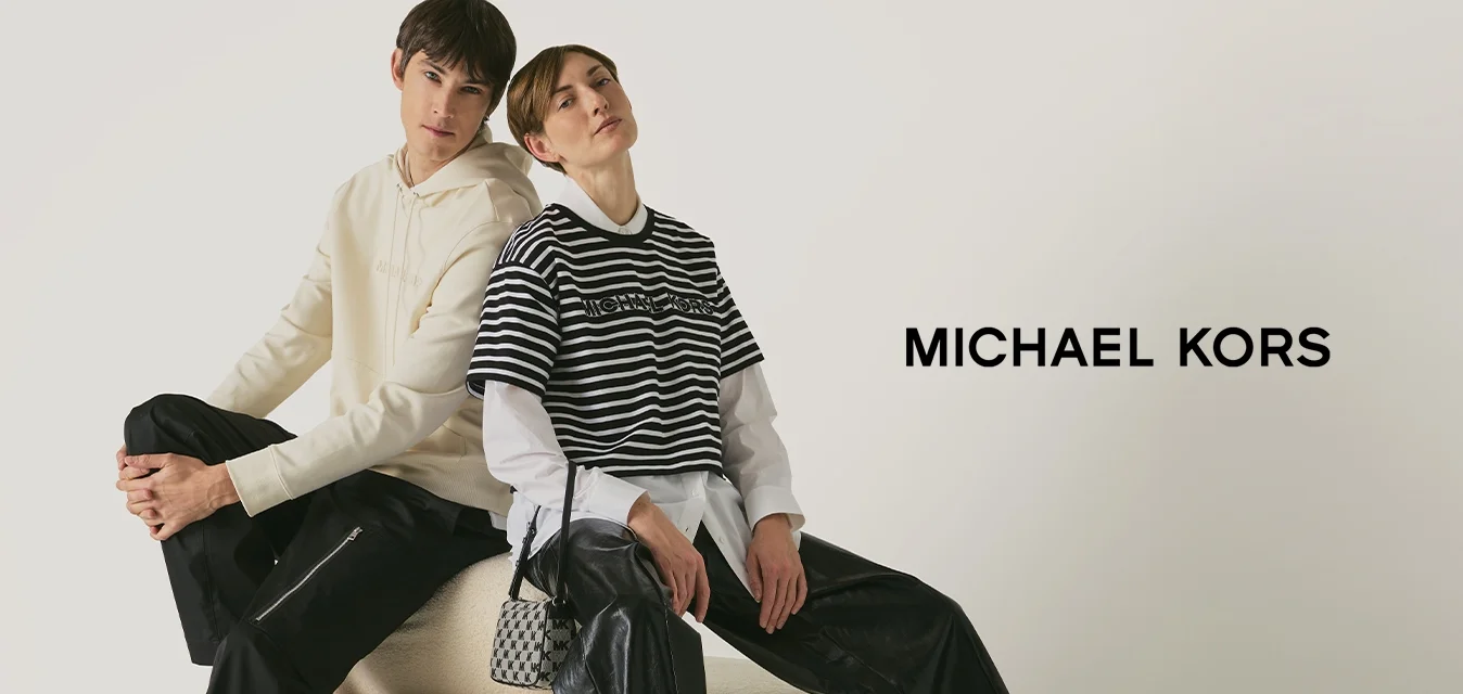 Twee jonge mensen zitten rug aan rug op een ronde beige poef, gekleed in casual chique outfits, tegen een witte achtergrond.