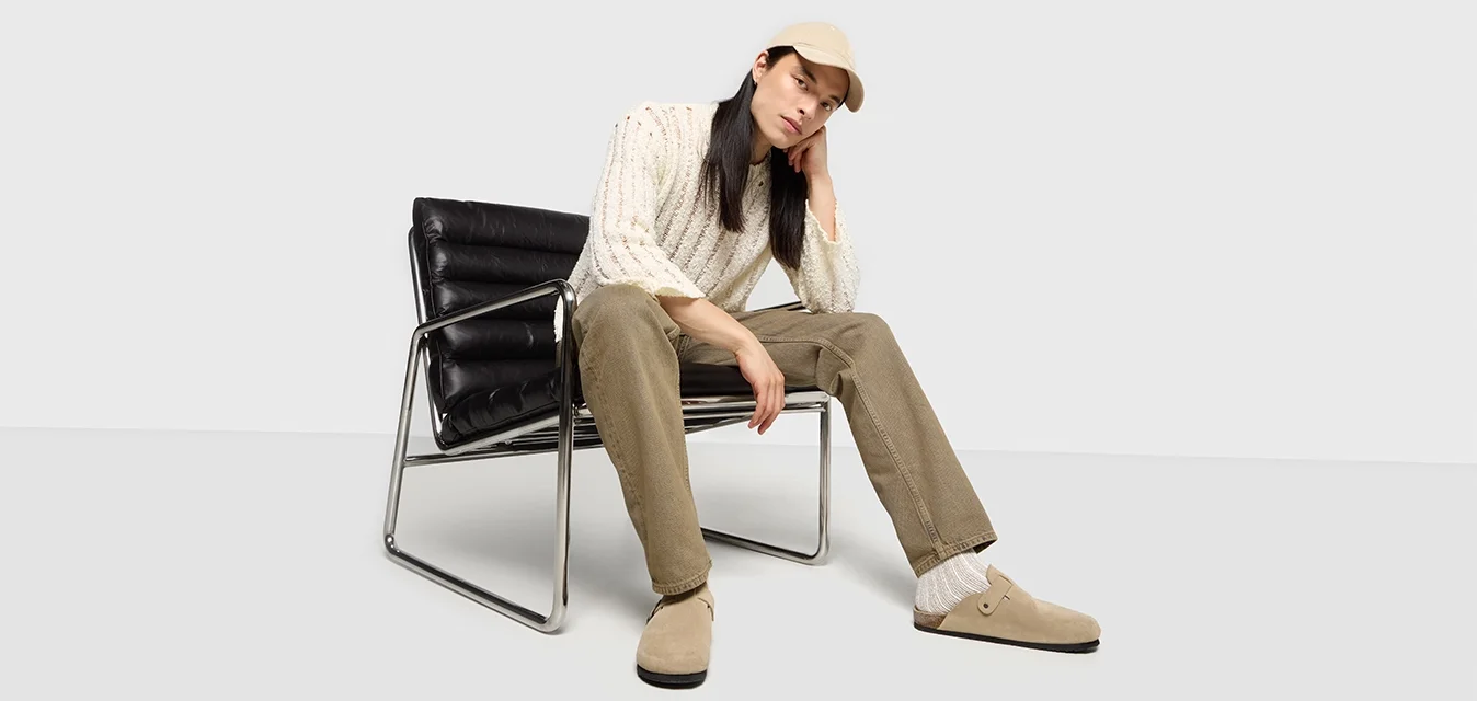 Zuecos de ante beige, pantalones de pana verde oliva, suéter de punto blanco, calcetines crema, sentado en una silla de cuero negro con marco de cromo.