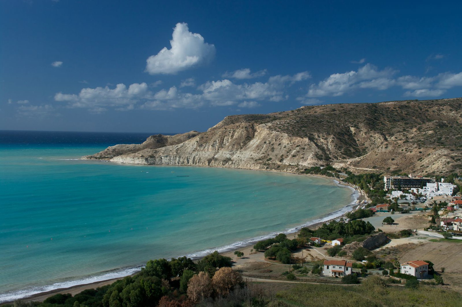 Aerial view of a turquoise bay with sandy beach, dramatic limestone cliffs and a small coastal village under a blue sky
