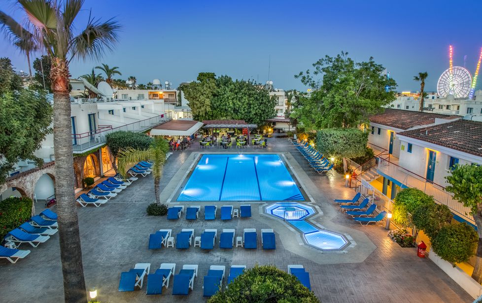Evening view of hotel swimming pool with rows of sun loungers, palm trees and lit ferris wheel on the skyline