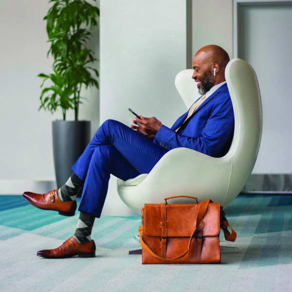 A well-dressed businessman in a blue suit sits in a contemporary white lounge chair, smiling while using his smartphone. He wears brown leather shoes and patterned socks, suggesting a stylish yet professional look. A tan leather briefcase rests on the floor beside him, with a green indoor plant in the background. The setting appears to be a bright, modern office or lobby with teal and gray carpeting.
