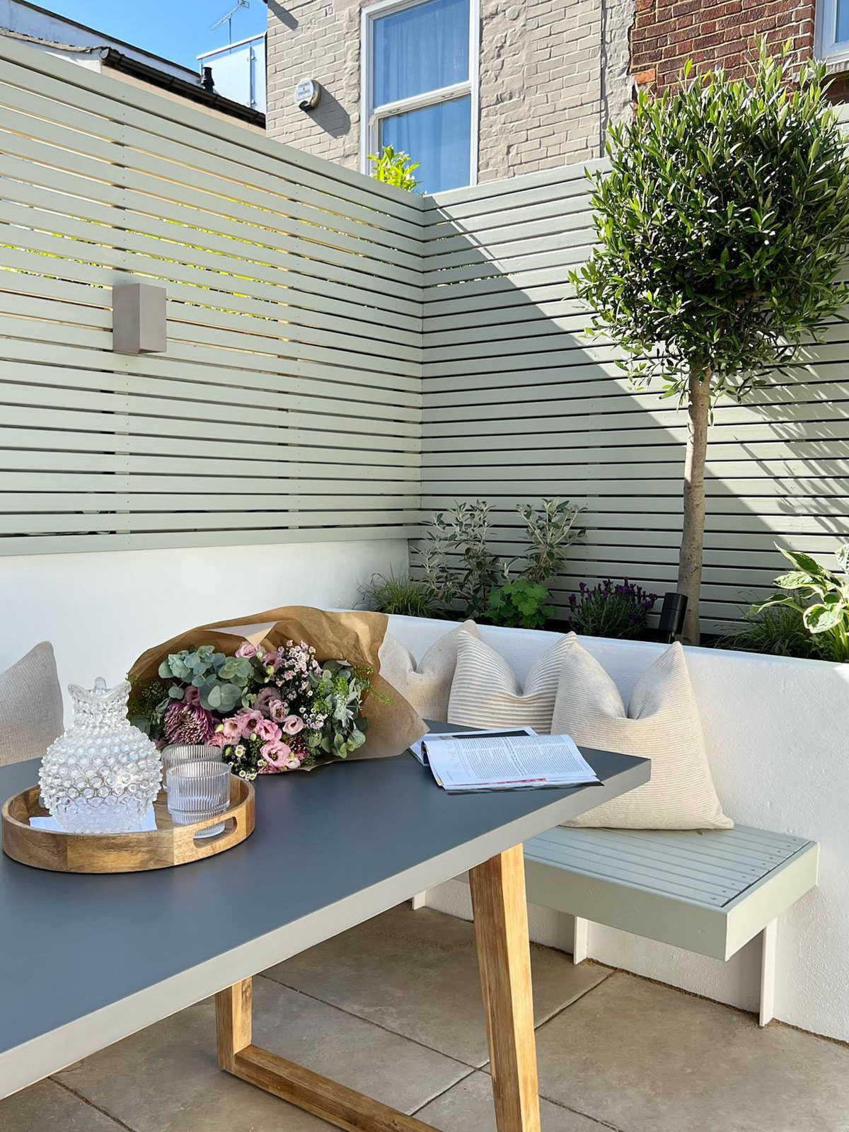 Modern patio with pale fencing, bench seating, and a table with flowers and glassware.