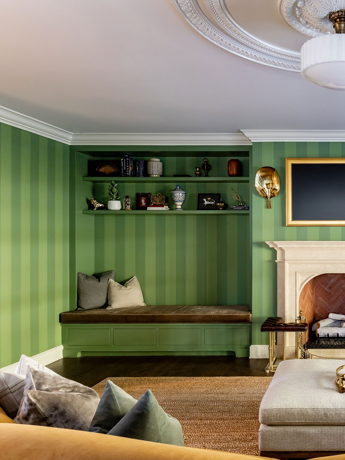 Bold green stripes dominate this delightful sitting room. Curl up on the built-in seat with its luscious velvet base and sink back onto the two plump cushions. Above you sit a curated collection of treasures and memories from books to vases and ornaments. At the edge of the image, we see part of a grand fireplace with a cream surround, the brushed gold candle sconce and mirror picking up the gold of the legs of the hearth accessories.