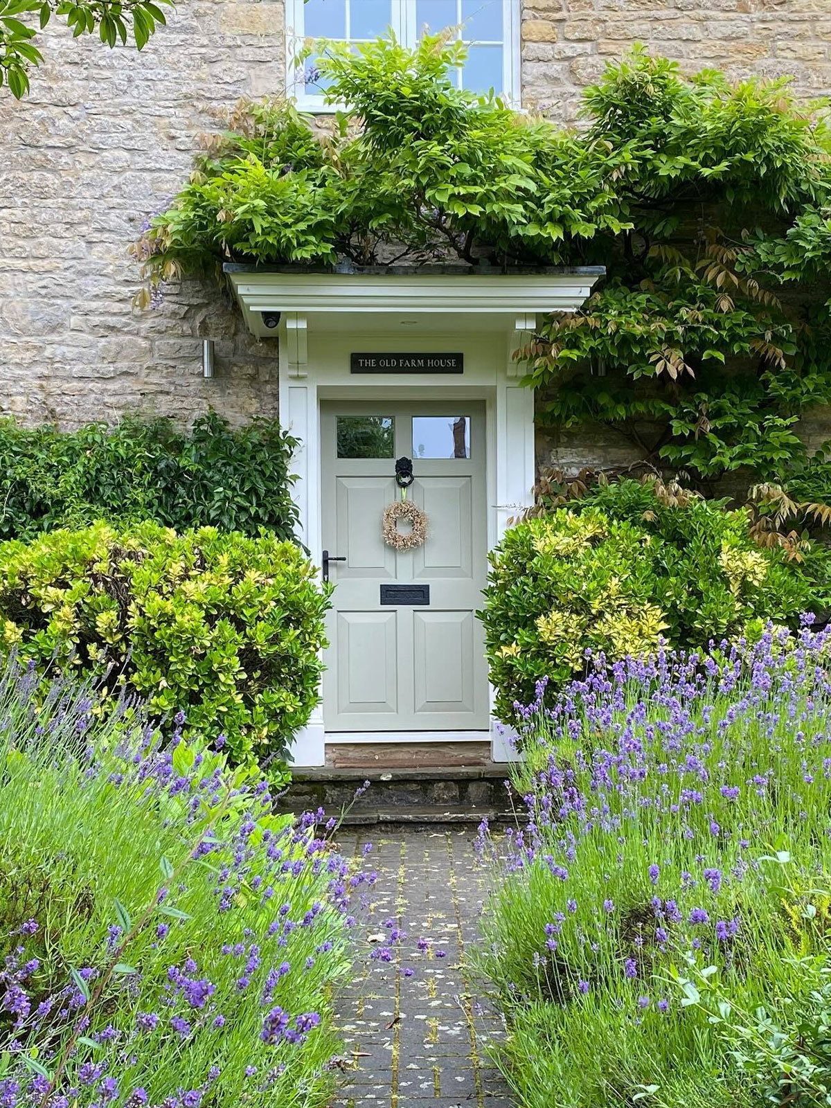 Front door of a stone farmhouse framed by green foliage and lavender pathway, with a wreath on the door.