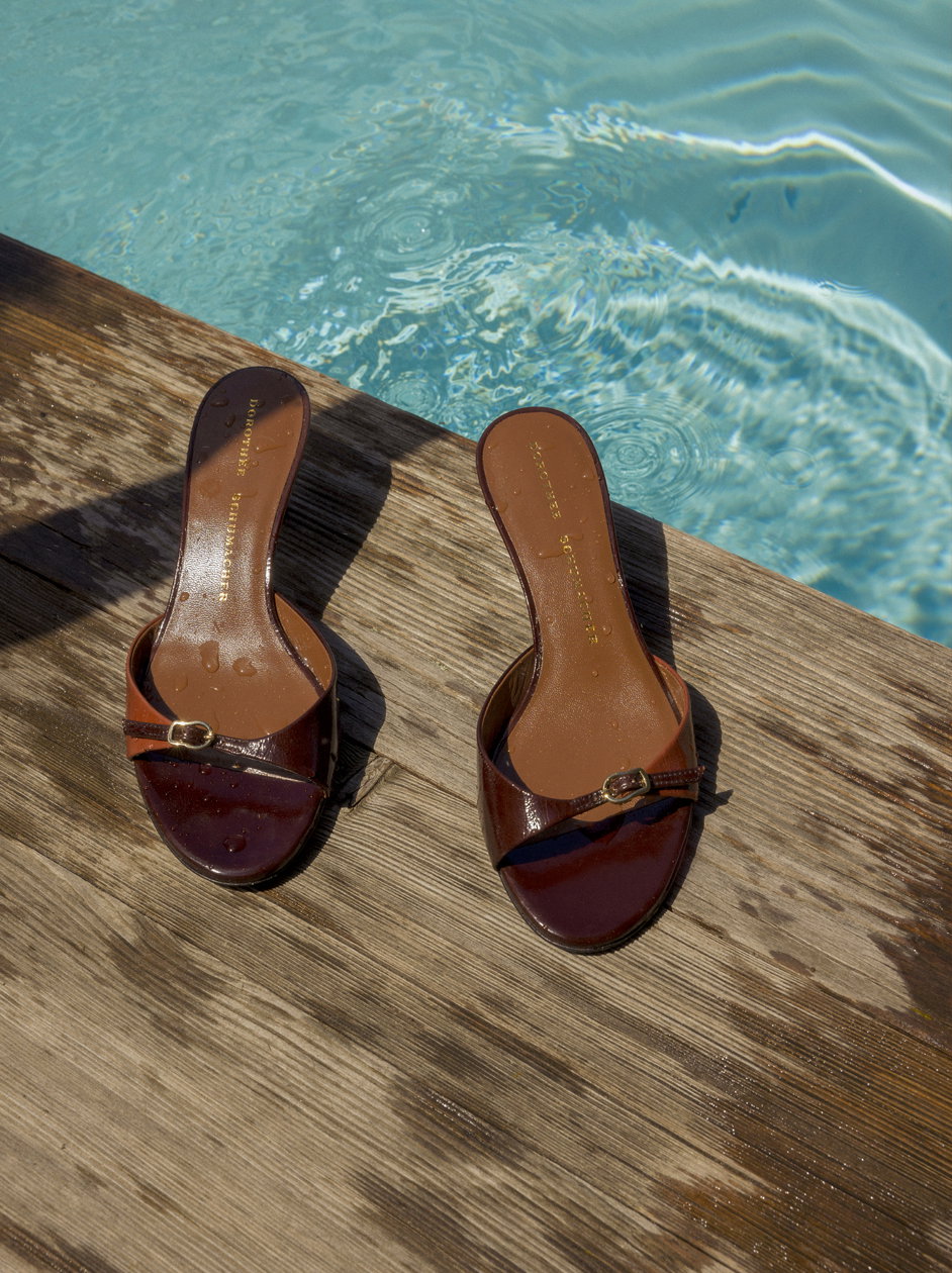 Ein Paar elegante braune Lackleder-Mules von DOROTHEE SCHUMACHER mit feinen goldfarbenen Schnallen stehen auf nassen Holzbohlen direkt am Rand eines glitzernden, blauen Pools.