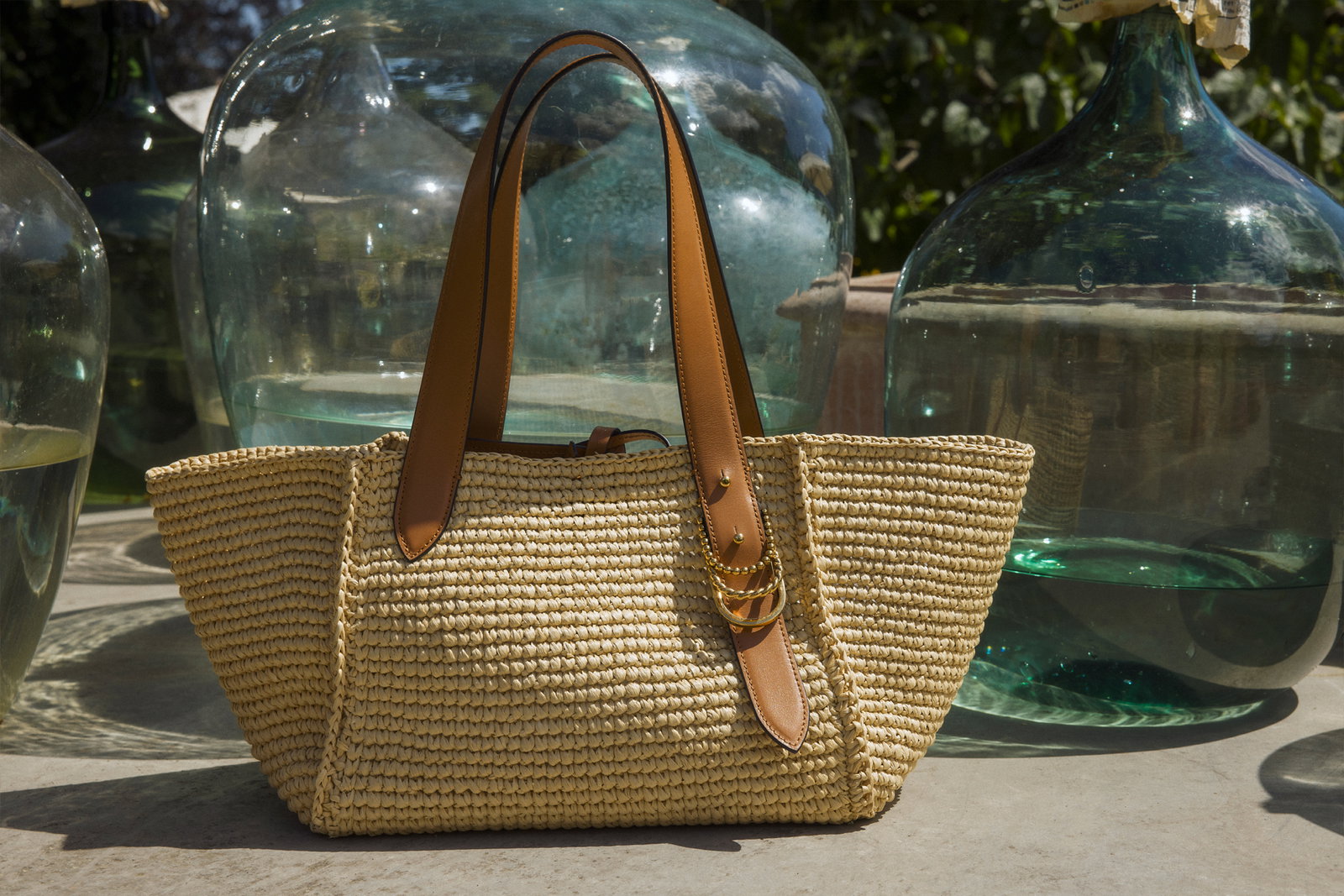 Die luxuriöse, handgehäkelte DOROTHEE SCHUMACHER Raffia Tote Bag präsentiert sich im Sonnenlicht vor großen, dekorativen Glasballons. Sie besticht durch geschmeidige braune Lederriemen und ein filigranes goldenes Kettendetail.