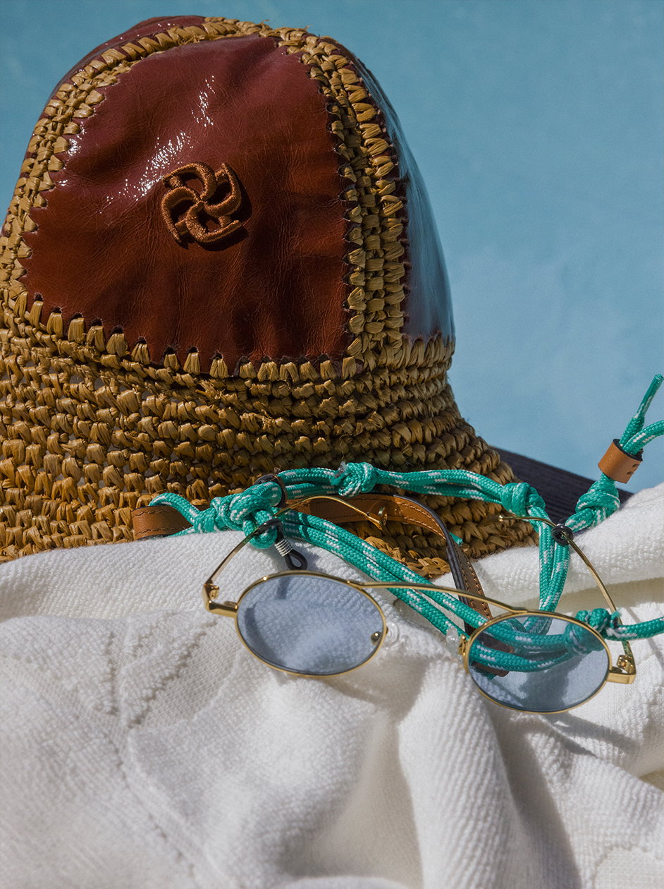 Ein sommerliches Stillleben von DOROTHEE SCHUMACHER am Pool: Ein Strohhut mit braunem Lederpatch und goldener Logo-Stickerei liegt neben einer runden, goldgefassten Sonnenbrille mit grüner Kordelkette auf einem weichen, weißen Handtuch.