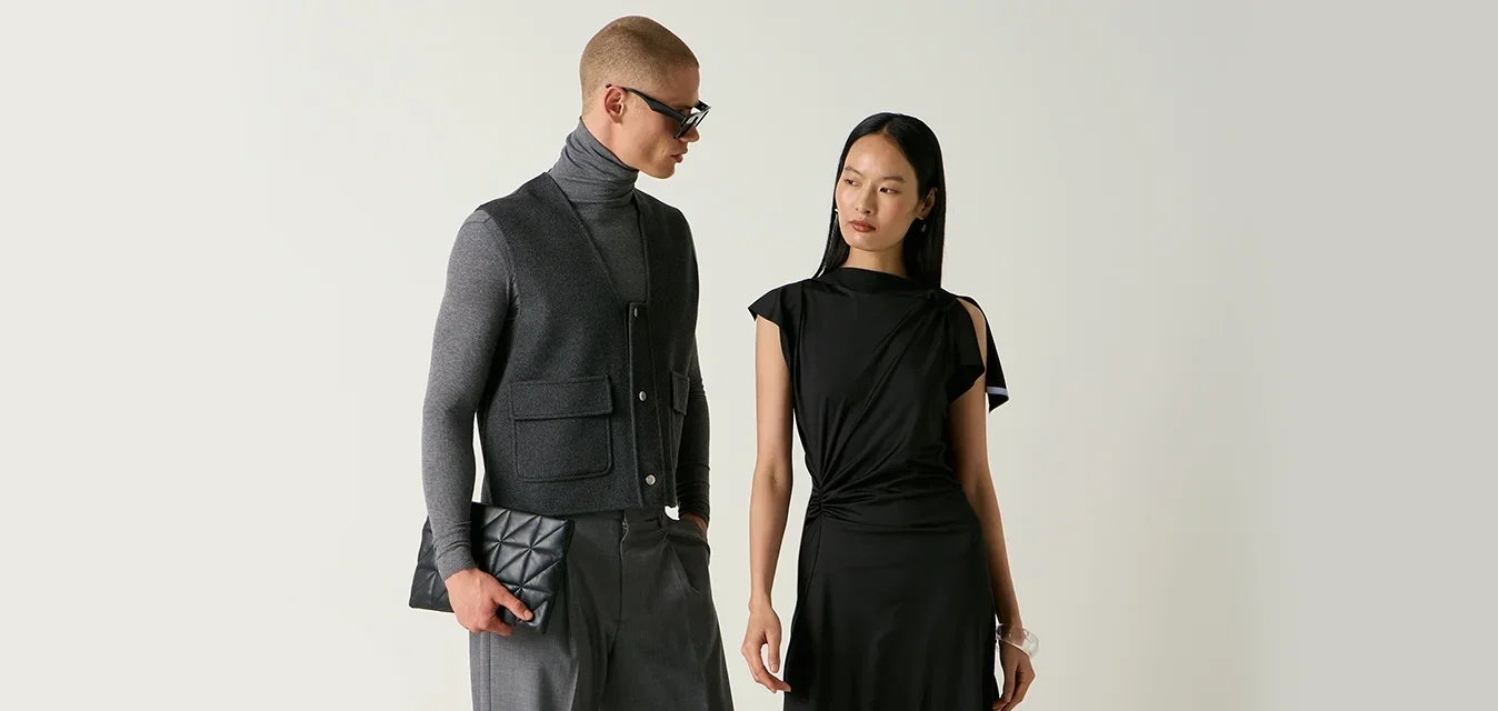 Homme en pull à col roulé gris, gilet et pantalon tenant une pochette, et femme en robe noire et chaussures debout près de chaises, dont une est renversée sur le sol.