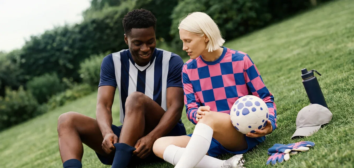 Deux joueurs de football sont assis sur l'herbe, se reposant ; l'un tient un ballon, avec des bouteilles d'eau, des gants et des casquettes à proximité dans un cadre de parc.