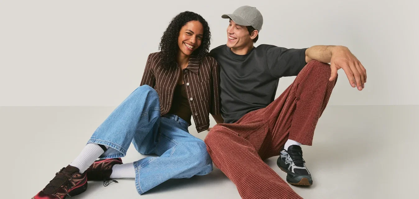 Dos jóvenes adultos sonriendo y sentados en el suelo, vistiendo ropa casual y zapatillas deportivas, frente a un fondo claro y liso.