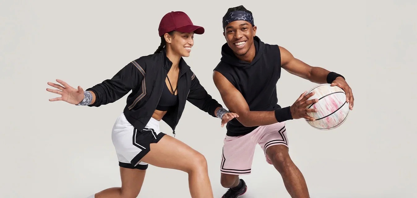 Hombre y mujer jugando al baloncesto, hombre botando el balón, mujer en posición defensiva, ambos vestidos con ropa deportiva y zapatillas.