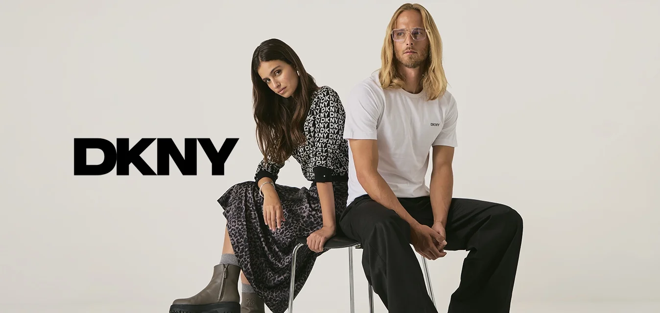 Una mujer con un vestido estampado y botas y un hombre con una camiseta blanca de DKNY y pantalones negros están sentados espalda con espalda en una silla contra un fondo liso.