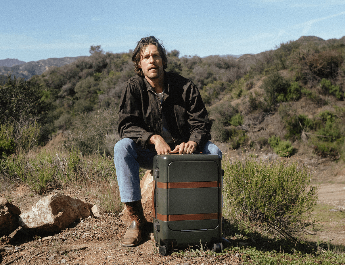A man sits on a rock in a hillside landscape, resting his hands on a green hardshell suitcase with brown leather straps.