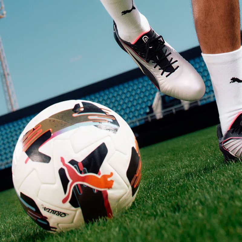 Close-up of footballer’s Puma boots kicking a white patterned match ball on green grass in a stadium