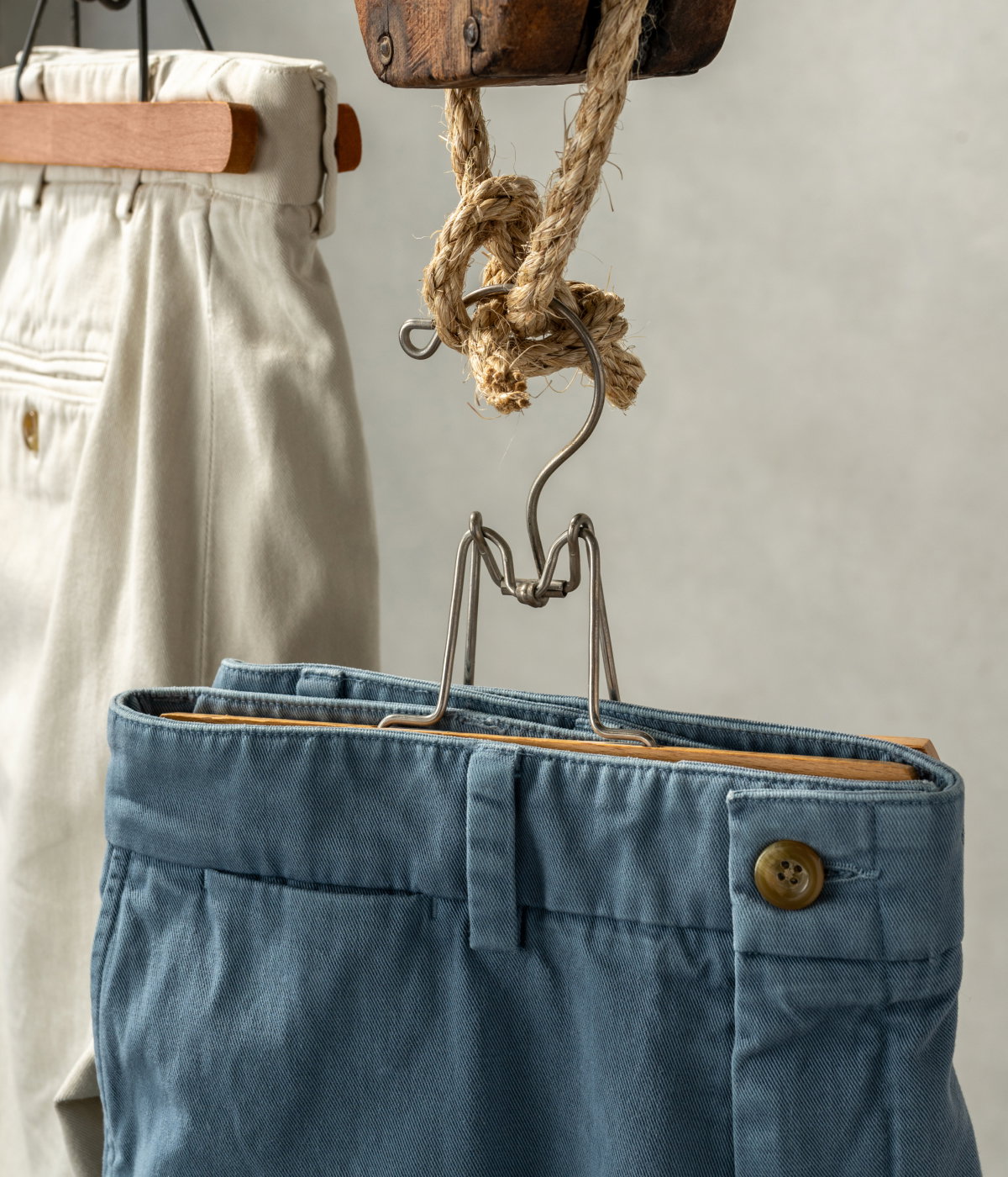 A blue pair of Bills Khakis shorts sits hanging from a vintage pulley, rope and hangar. Another pair of bone colored shorts hangs the same in the background