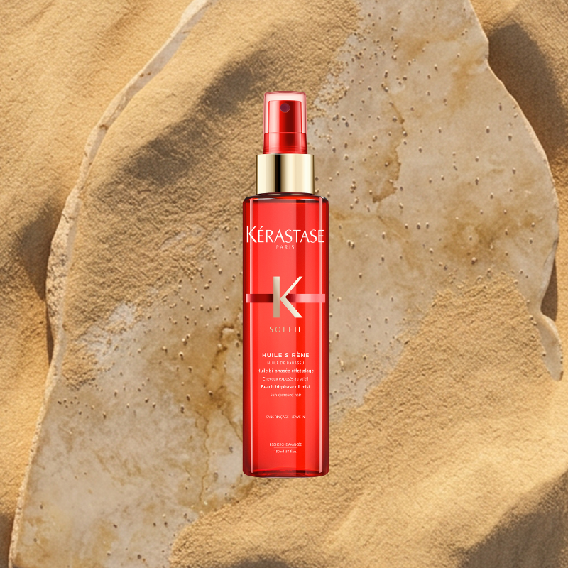 Flacone spray rosso Kérastase Soleil Huile Sirène appoggiato su roccia e sabbia