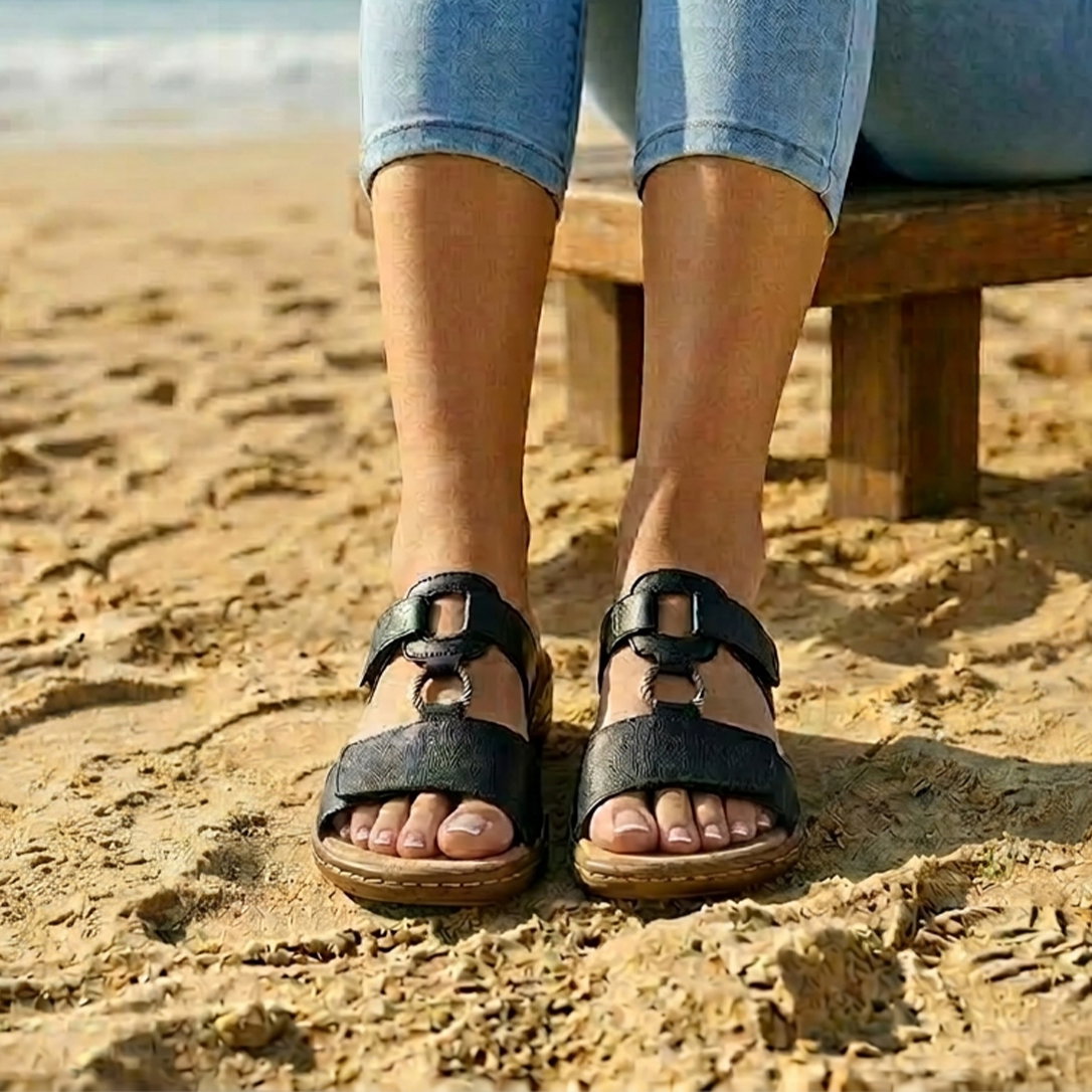 Ein Model sitzt am Strand auf einer Holzbank und trägt schwarze Sandalen und eine helle Jeans.
