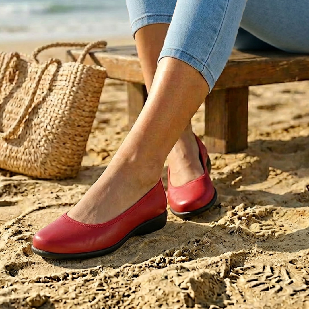 Ein Model sitzt am Strand auf einer Holzbank, trägt rote Ballerinas und eine helle Jeans. Links liegt eine geflochtene Tasche.