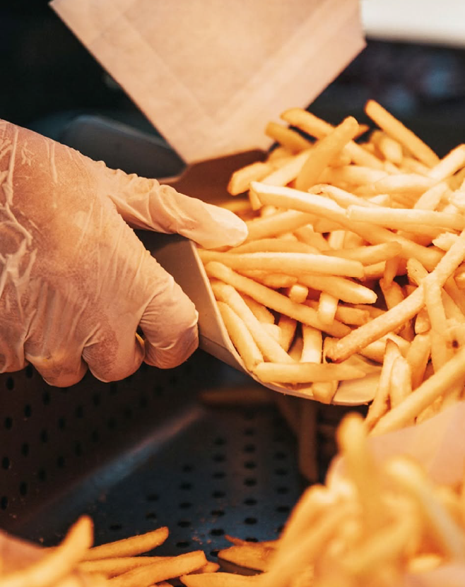 French fries being scooped into a serving container from a heated holding station in a commercial foodservice setting.