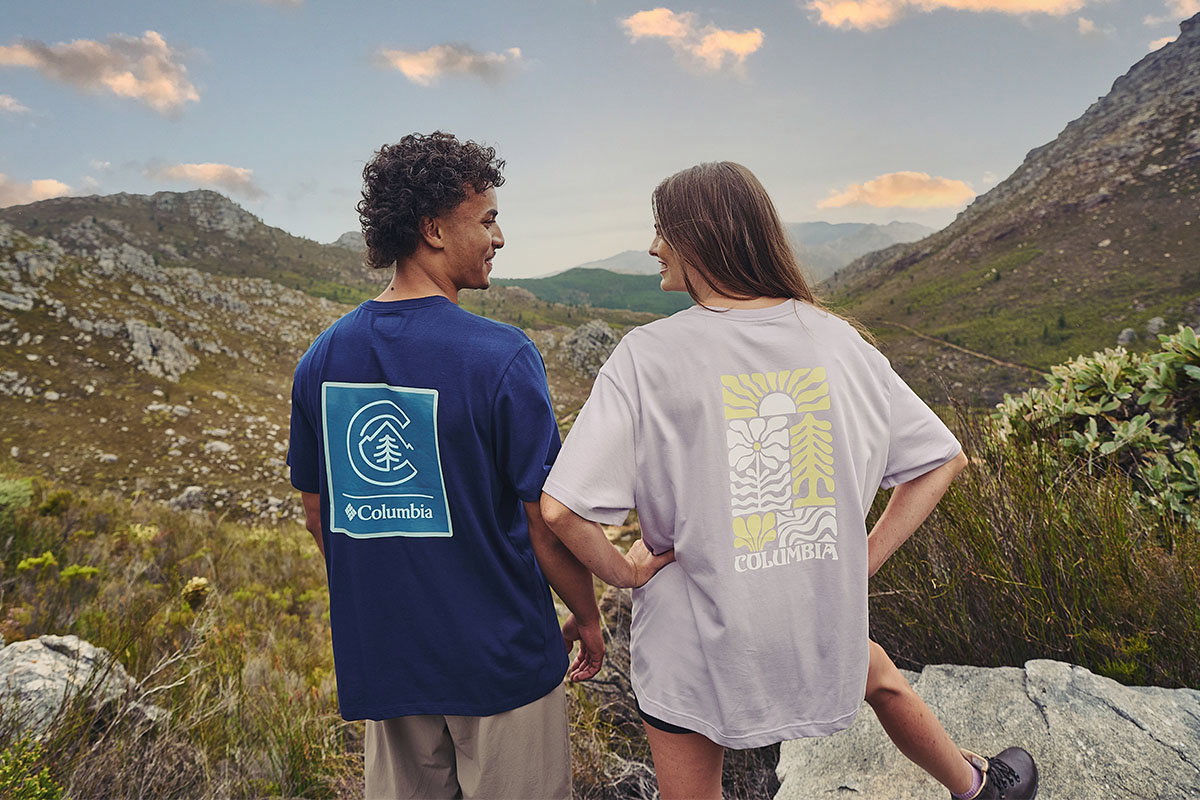 Two people stand on a rocky mountain hillside, viewed from behind, wearing oversized Columbia graphic T-shirts. One T-shirt is navy blue with a large blue-and-white logo graphic on the back, and the other is light grey with a yellow-and-white nature-inspired graphic. Rolling hills and mountains stretch into the background beneath a partly cloudy sky. The outdoor scene focuses on casual apparel in a scenic hiking landscape.
