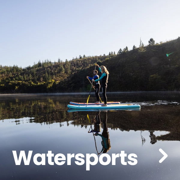 Two people stand on paddleboards in the middle of a calm lake, using paddles as they glide across still water that reflects the surrounding landscape. Forested hills line the far shore under a clear sky. Large white text reading “Watersports” appears across the bottom of the image, and a right-arrow navigation icon is displayed on the right side. The scene is bright and outdoors, with clear reflections visible on the water’s surface.